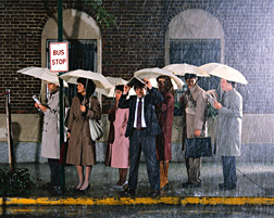 Photo: Rain falling on an urban street and people standing at a bus stop.